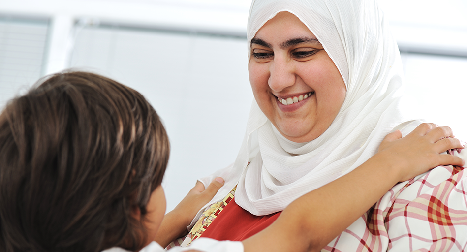 A smiling woman looks at a child who has their arms around her.