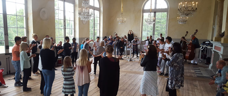 Musiker spelar i en stor sal med trägolv. Det står folk runtomkring och applåderar.