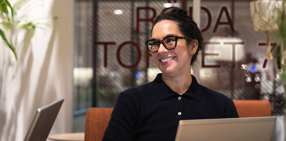 Kvinna med glasögon som sitter vid en laptop och ler.