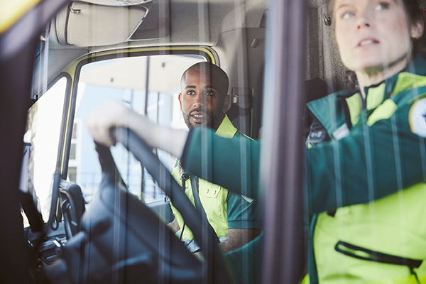 Två sjukvårdare sitter i ambulansen. Kvinnan kör och mannen tittar ut genom fönstret. Bilden är tagen från sidan i dagsljus.