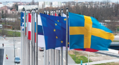 Flera nationsflaggor, däribland EU-flaggan, vajar på flaggstänger utomhus med Sveriges flagga i förgrunden.